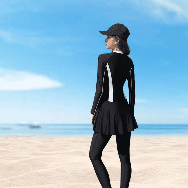 A person wearing a black swim suit with a skirt, standing on a beach under a blue sky.