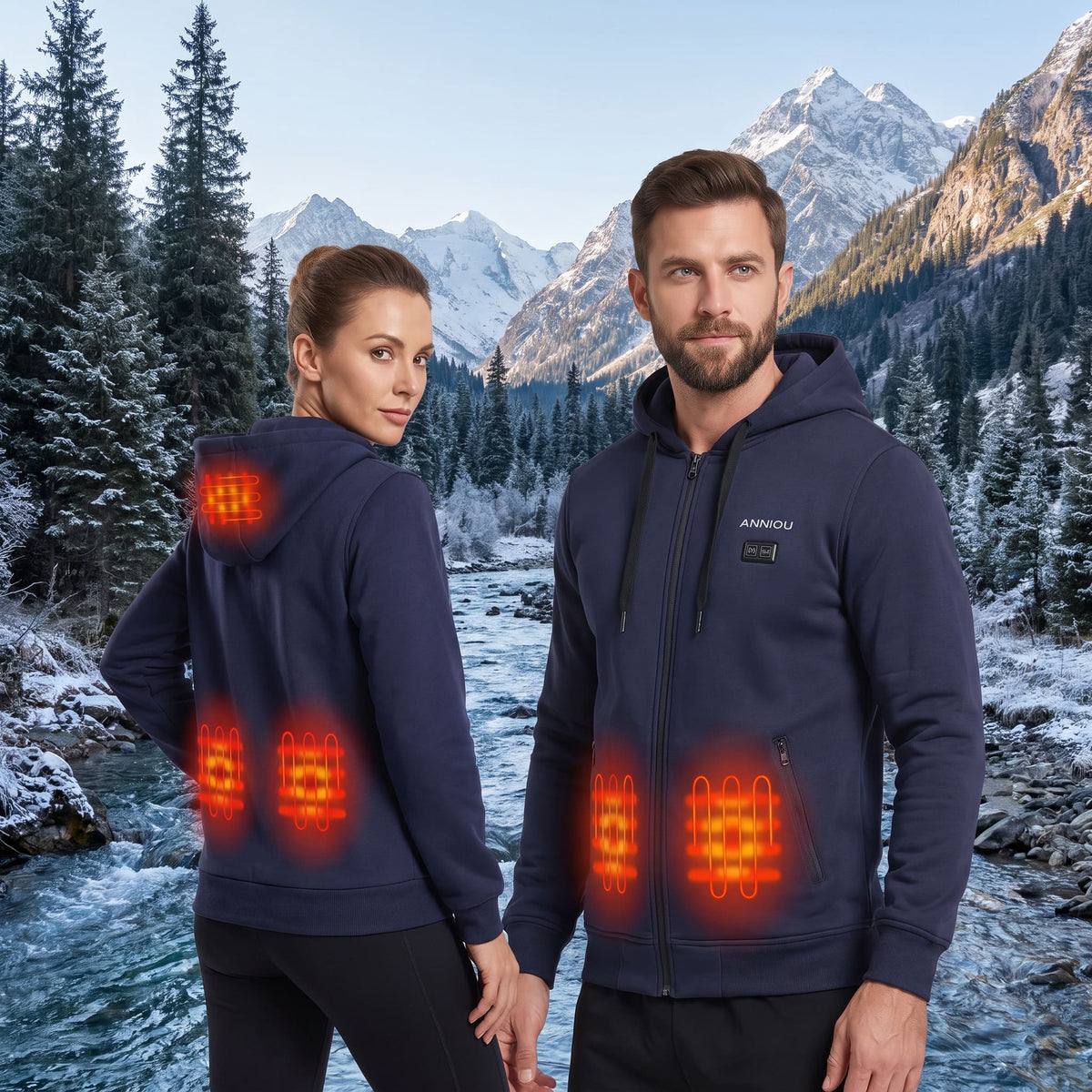 A man and a woman wearing navy blue heated hoodies stand in a snowy mountain landscape by a river. The hoodies have glowing orange heat-map graphics showing heated zones on the back and front torso.