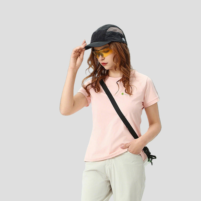Woman in a pink Anniou t-shirt and cap standing in front of a scenic mountain landscape with a small building and vehicle.