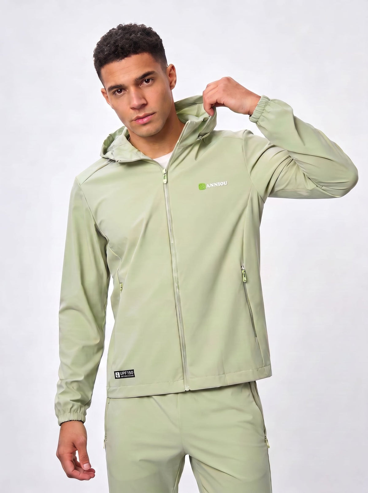 A man wearing a light sage green hooded jacket and matching pants, posing against a light gray studio background. The jacket features a small green and white logo on the left chest and a black brand patch near the hem.