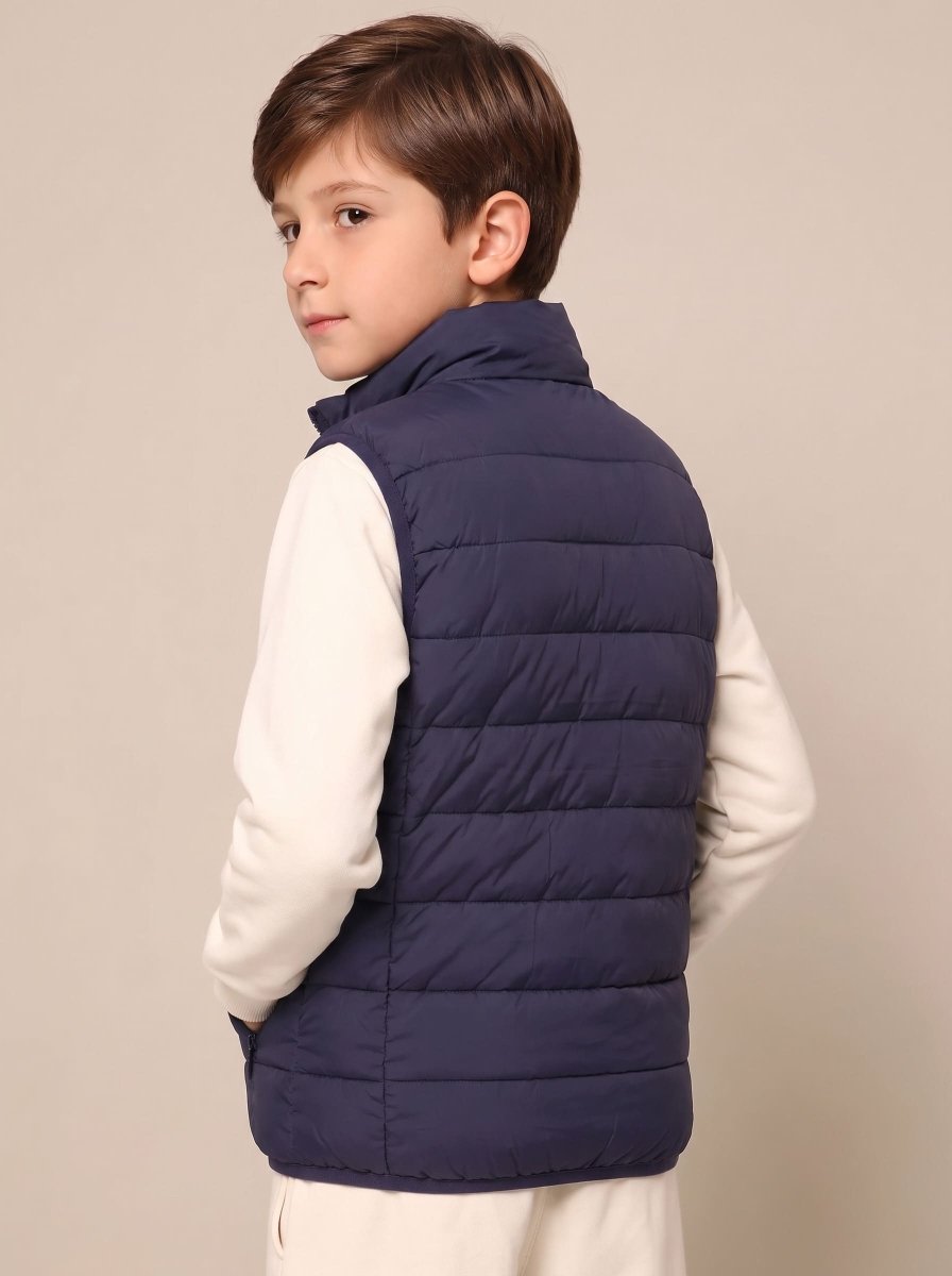 A young boy wearing a navy blue quilted puffer vest over a cream-colored long-sleeve shirt, photographed from the back against a solid light beige background.
