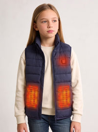 A young girl wearing a navy blue quilted heated puffer vest over a cream sweatshirt and jeans, with glowing orange heat-map graphics highlighting the heating zones.