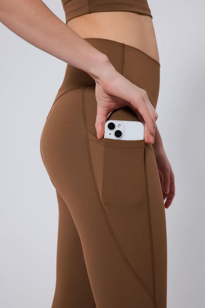 Person wearing brown leggings with a pocket holding a phone on a plain background