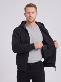 Male model showing inner battery pocket of black heated hooded jacket, unzipped coat, light gray studio background, commercial apparel photo