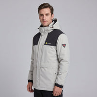 Studio portrait of European male model in #ivorywhite men's heated hooded jacket, wind-resistant thermal outdoor winter coat with brand logo and arm patch, solid light grey background, apparel product photography
