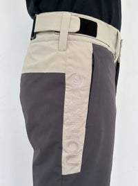 Close-up of a person wearing color-blocked ski pants with a beige upper section, dark gray lower section, and a vertical beige panel on the leg featuring the word 'ANNIOU' in raised embroidery.