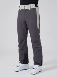 Dark gray ski pants with beige side panels and a matching beige waistband, shown on a model wearing white ski boots against a light gray studio background.
