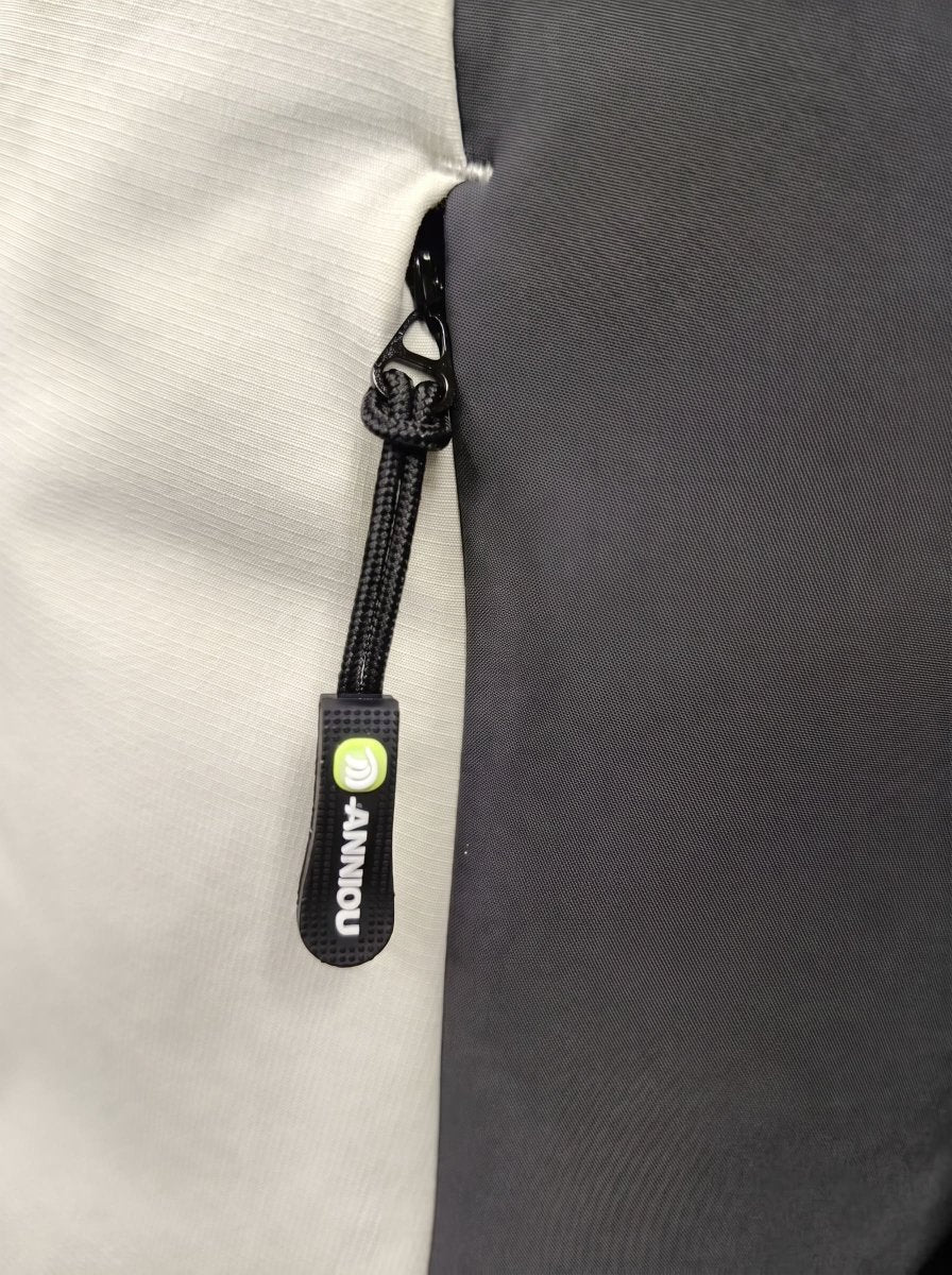 Close-up of a black zipper pull tab with a bright green circular logo and white vertical text reading 'ANNIOU' on a light gray fabric next to a dark gray panel.