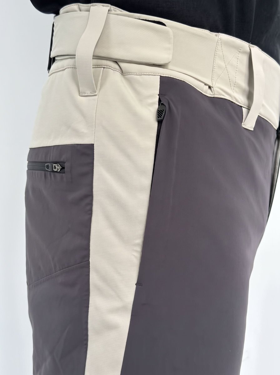Close-up of a pair of color-blocked snowboard pants with a light beige waistband and side panel, a dark gray vertical stripe, and a dark gray zippered pocket on the thigh.