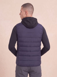 A man viewed from the back wearing a dark navy quilted puffer vest over a black long-sleeve shirt and gray pants, set against a plain beige background.