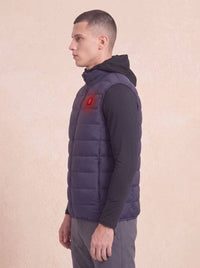 A man in profile wearing a dark navy quilted puffer vest over a black long-sleeve shirt and gray pants, standing against a textured beige background. A red glow effect is visible on the chest of the vest.