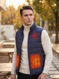 A man wearing a navy blue quilted puffer vest over a cream turtleneck sweater, standing outdoors in a softly blurred park setting. The vest has glowing orange heat-map overlays highlighting its front heating zones.