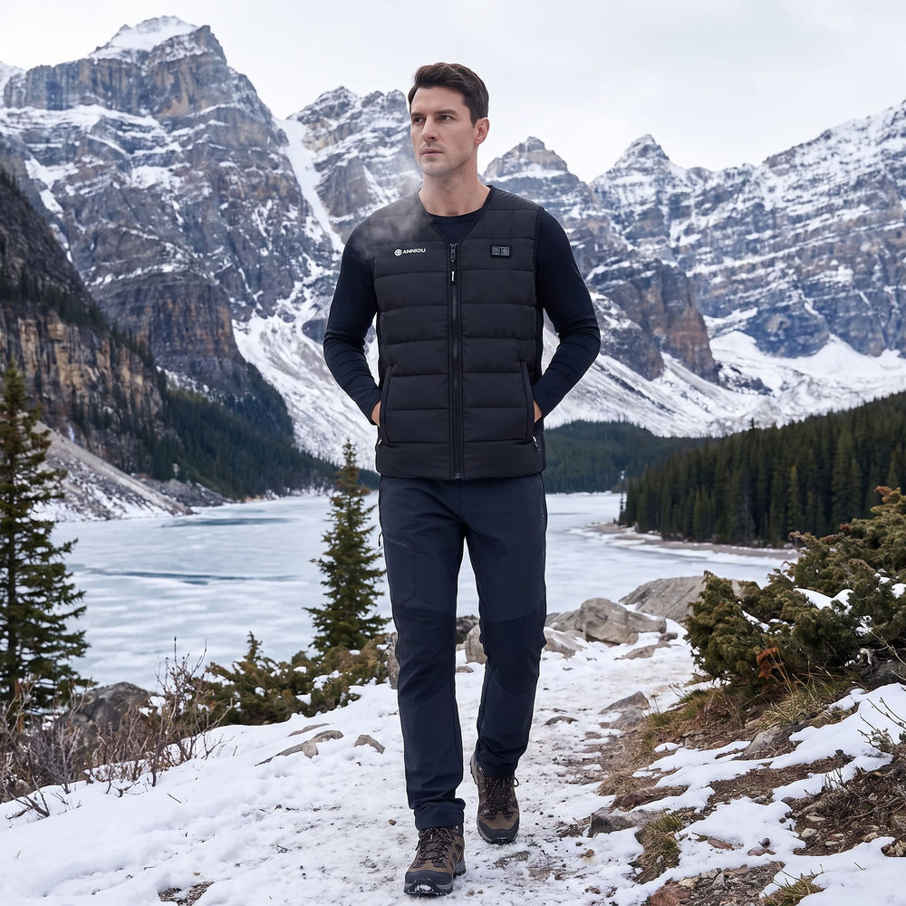 A man wearing a heated black puffer vest over a black long-sleeve shirt and dark pants stands on a snowy mountain path with a frozen lake and snow-covered peaks in the background.