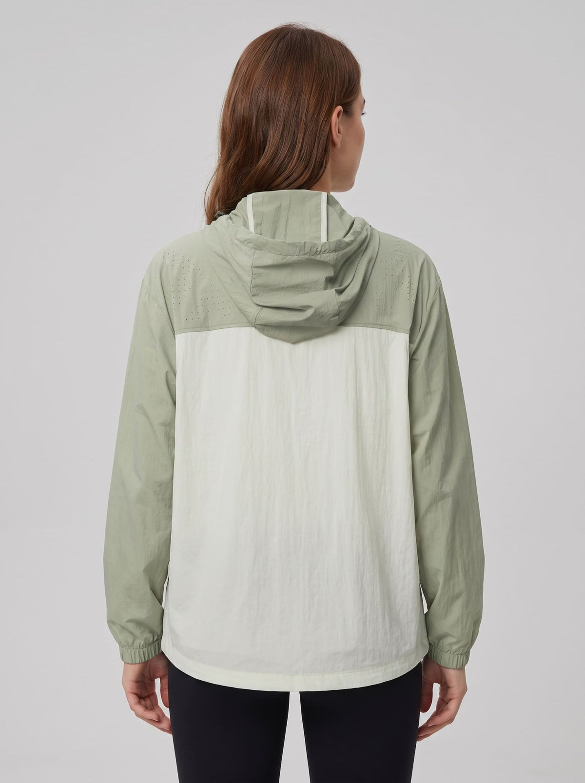A woman viewed from the back wearing a lightweight hooded jacket with a sage green upper section and sleeves, and an off-white lower back panel, against a plain light gray studio background.
