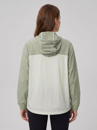 A woman viewed from the back wearing a lightweight hooded jacket with a sage green upper section and sleeves, and an off-white lower back panel, against a plain light gray studio background.