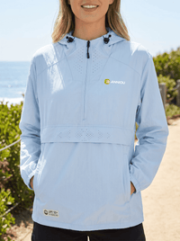 A woman wearing a light blue hooded windbreaker jacket with a half-zip front, drawstrings, and a visible Annou logo on the left chest, photographed outdoors near the beach.