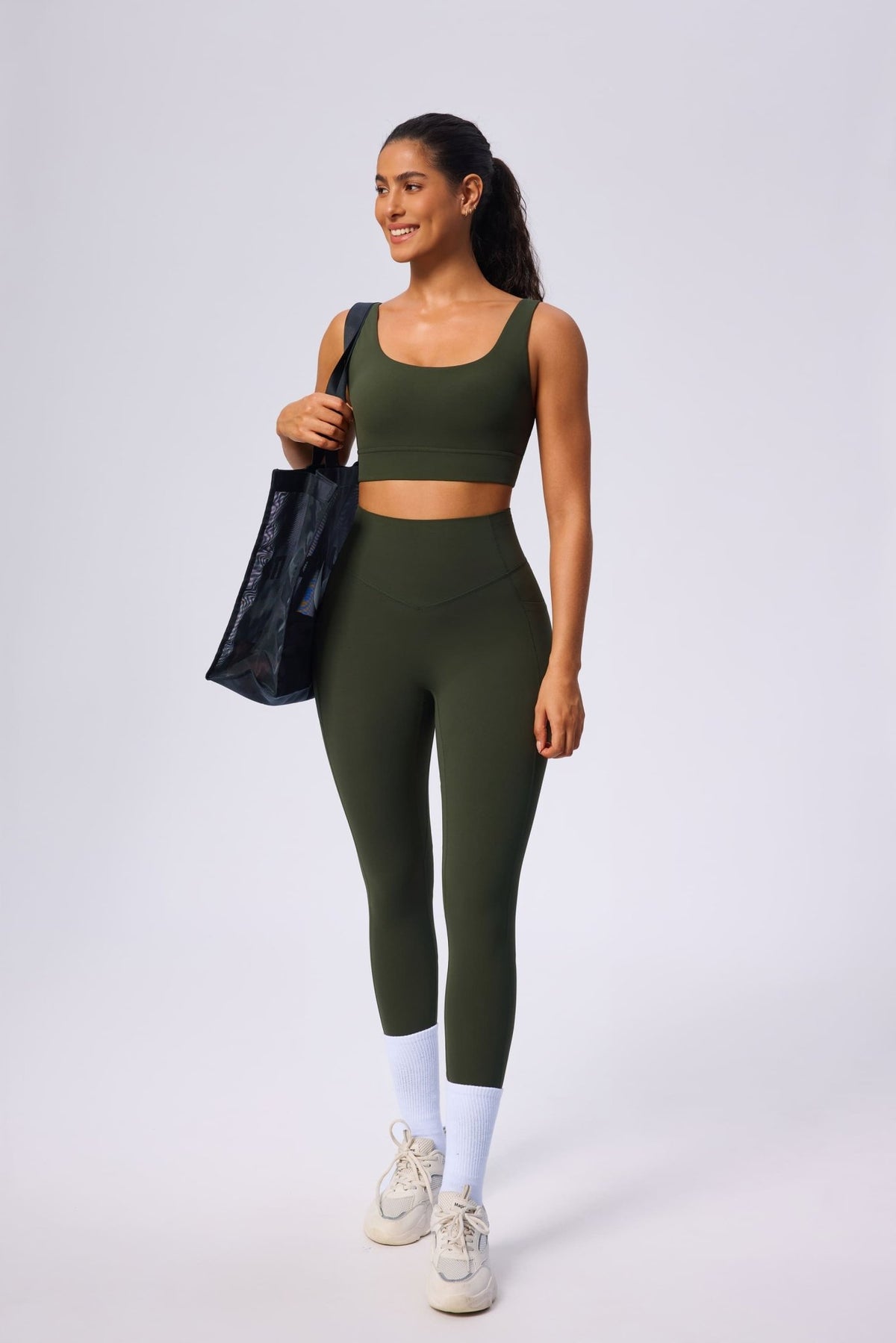 Woman wearing an olive green athletic outfit with leggings and a sports bra, holding a black bag on a white background.