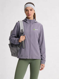 A woman modeling a light purple hooded athletic jacket with a green and white Anniou logo on the left chest, paired with olive green leggings and a gray backpack against a light gray studio background.