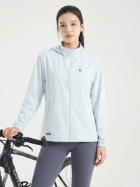 A woman wearing a fogblue hooded athletic jacket and dark gray leggings stands next to a black bicycle against a plain light gray studio background.