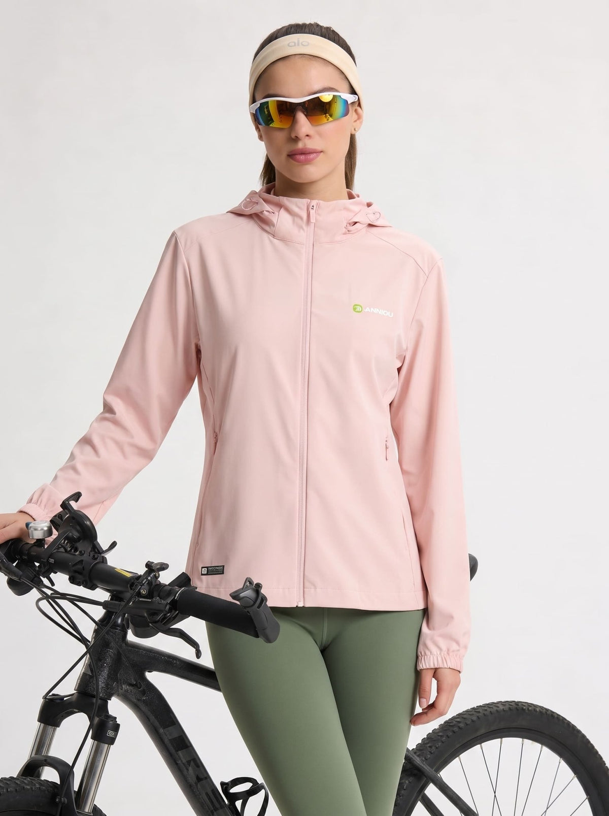 A woman wearing a lightpink hooded athletic jacket, olive green leggings, a beige headband, and mirrored sunglasses stands next to a black bicycle against a light gray studio background.