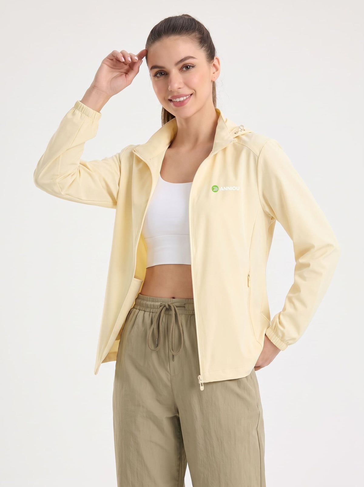 A woman modeling a lightyellow lightweight zip-up jacket over a white sports bra and olive green drawstring pants, photographed against a plain light gray studio background.