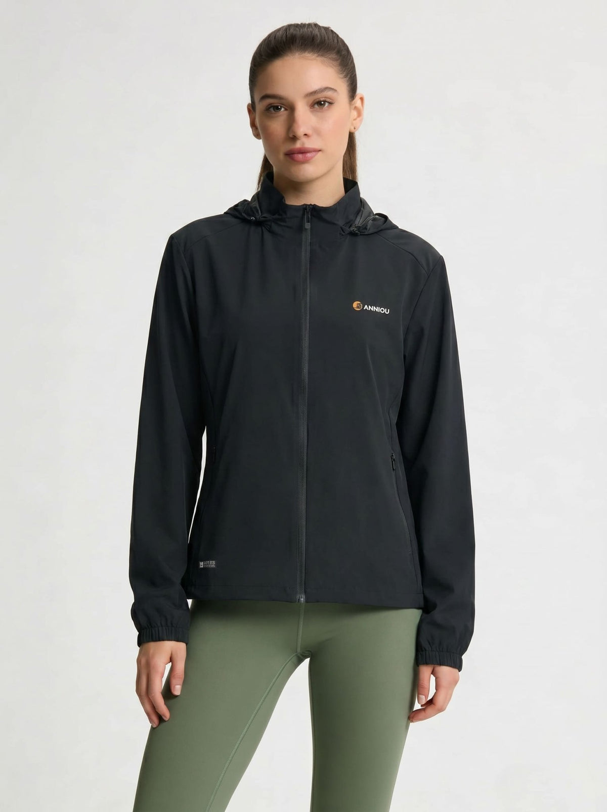 A woman modeling a black hooded zip-up jacket with a small orange and white Anniou logo on the left chest, paired with olive green leggings, against a light gray studio background.