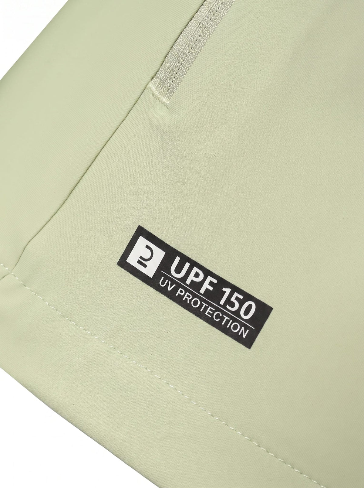Close-up of a light sage green fabric item showing a black rectangular label that reads 'UPF 150 UV PROTECTION' near a zipper.