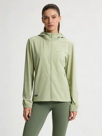 A woman modeling a lightgreen hooded athletic jacket with a full zipper and matching sage green leggings against a plain light gray studio background.