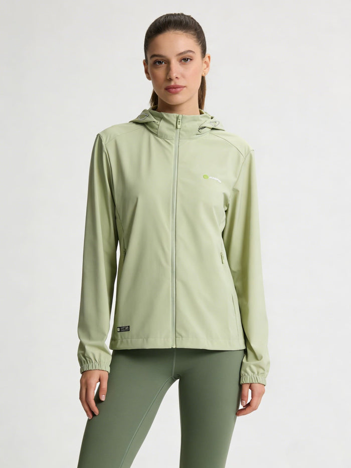 A woman modeling a lightgreen hooded athletic jacket with a full zipper and matching sage green leggings against a plain light gray studio background.