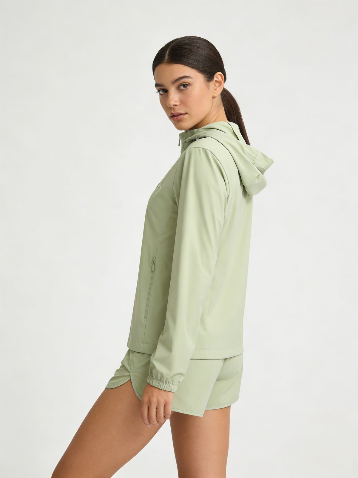 A woman modeling a light sage green hooded jacket and matching shorts against a plain light gray studio background.
