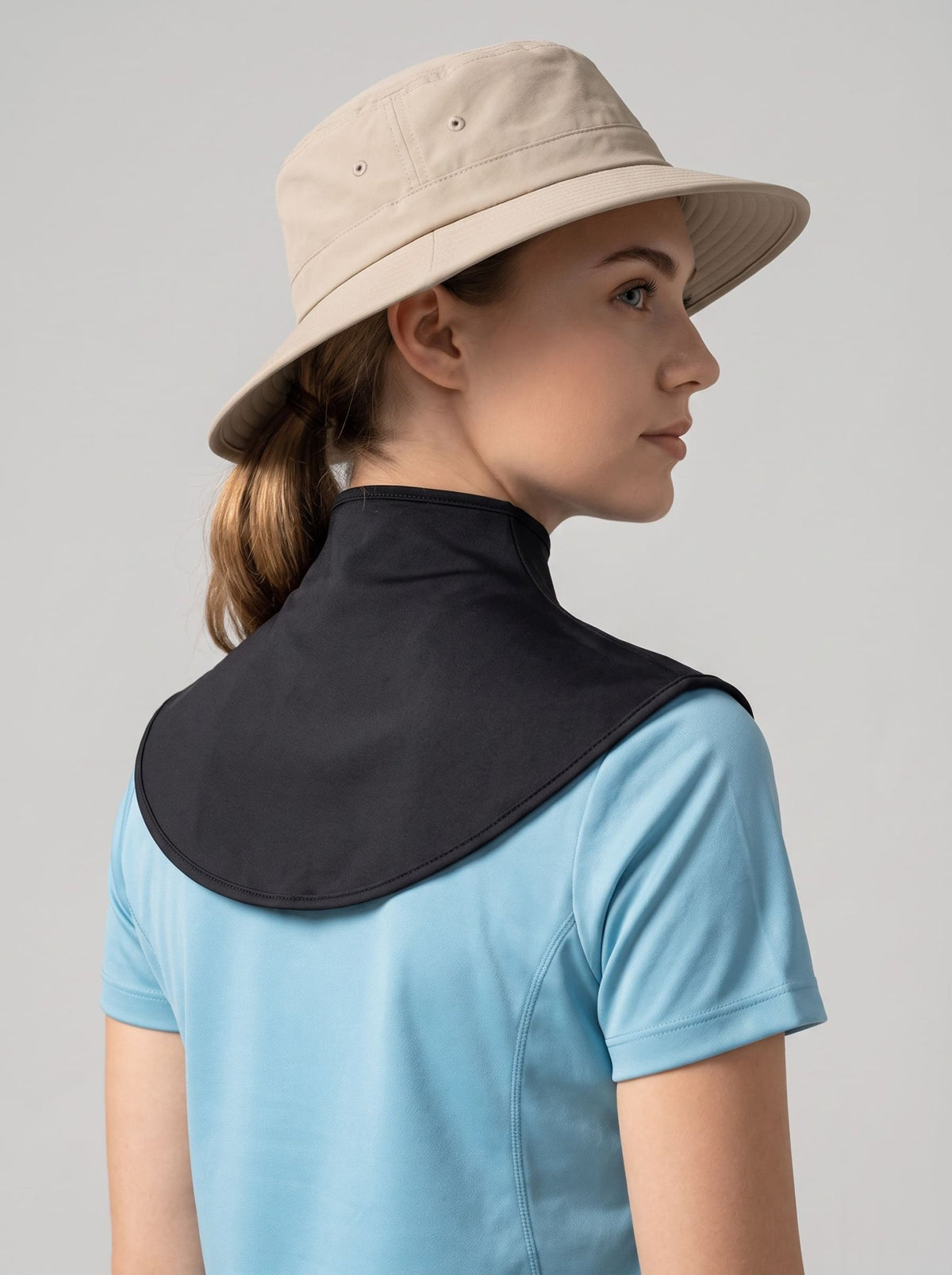 A woman shown from the back wearing a light beige bucket hat and a light blue short-sleeve shirt, with a black neck shield attached behind her neck, against a plain light gray studio background.