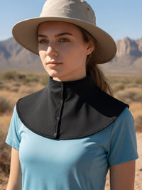 A woman wearing a light beige wide-brim sun hat and a light blue short-sleeve shirt, with a black high-neck collar bib accessory around her neck, photographed outdoors in a desert landscape.