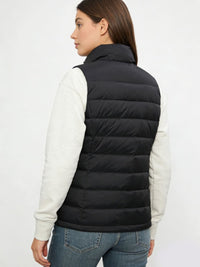 A woman shown from the back wearing a black puffer vest over a light heather gray long-sleeve top and blue jeans, against a plain light gray studio background.