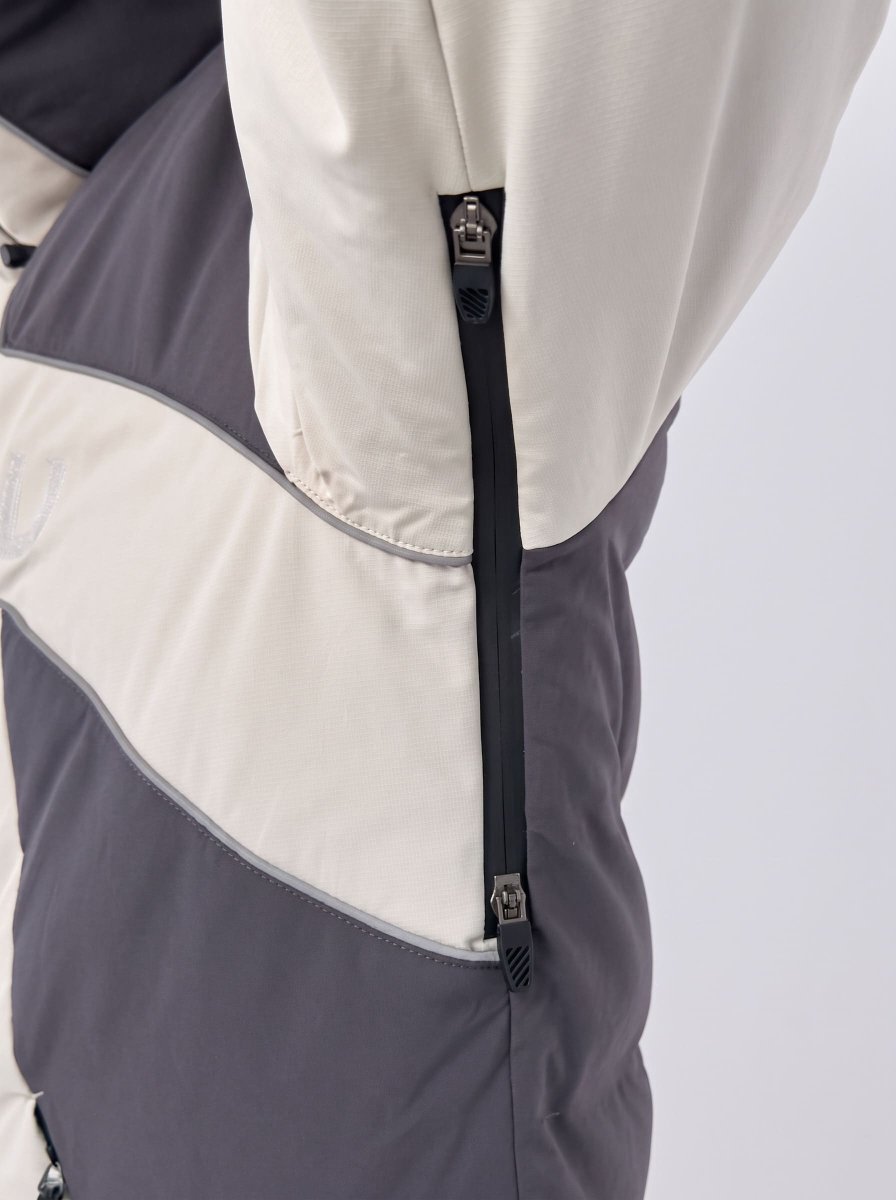 Close-up of a color-blocked heated ski jacket showing a diagonal split between off-white and dark gray panels, with a black zipper running vertically for beathability