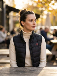 A woman wearing a black quilted puffer vest over a cream turtleneck sweater, sitting at an outdoor cafe table with a warm, blurred background.