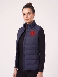 A woman wearing a navy blue heated puffer vest over a black long-sleeve top, shown against a plain light background. A glowing red logo on the chest indicates the brand Heatone.