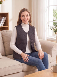 Woman sitting on a beige sofa, wearing a #black snap‑button heated gilet over a cream turtleneck sweater and blue jeans, bright cozy living‑room setting.